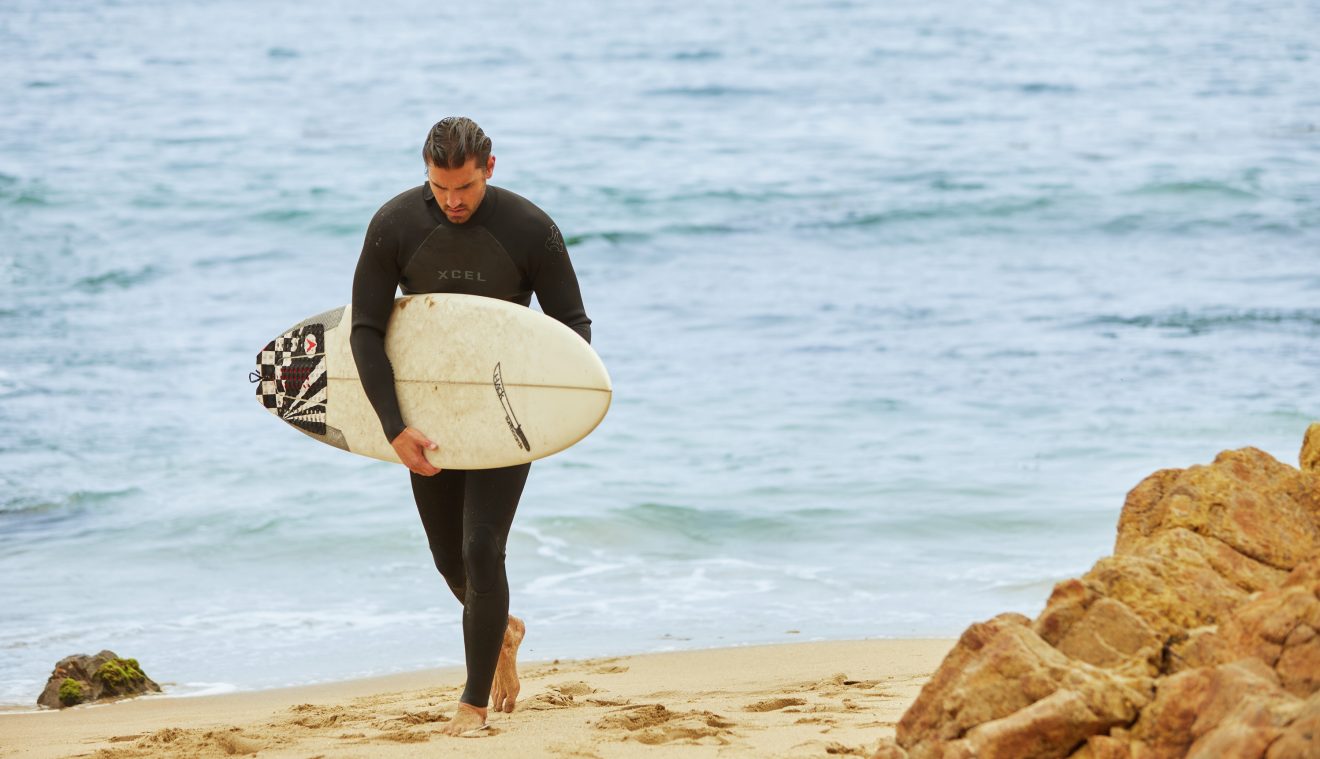 Exterior view of man walking on the beach holding a surfboard.